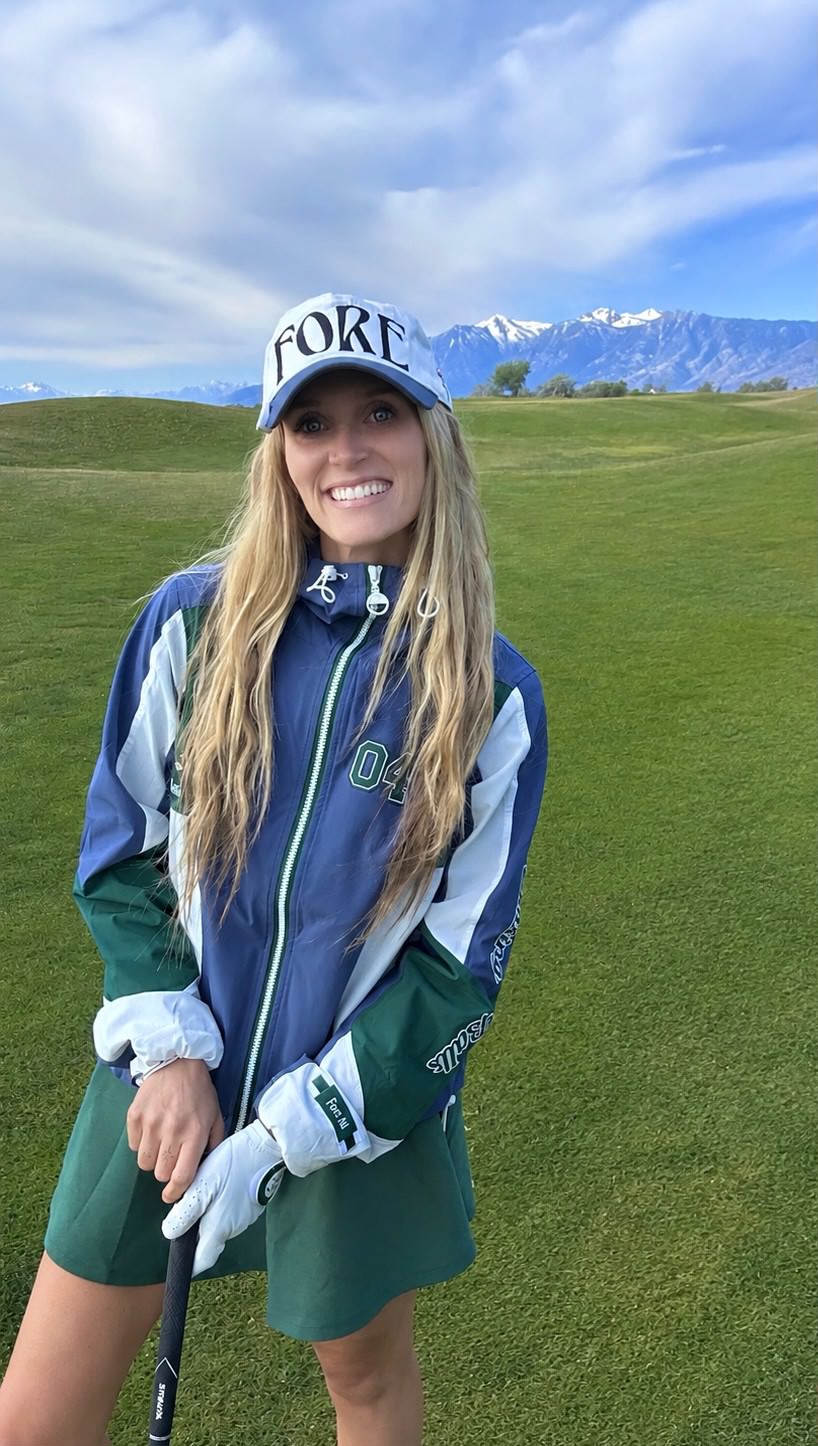 Kristen Catullo on the golf course holding a club, in a FORE cap